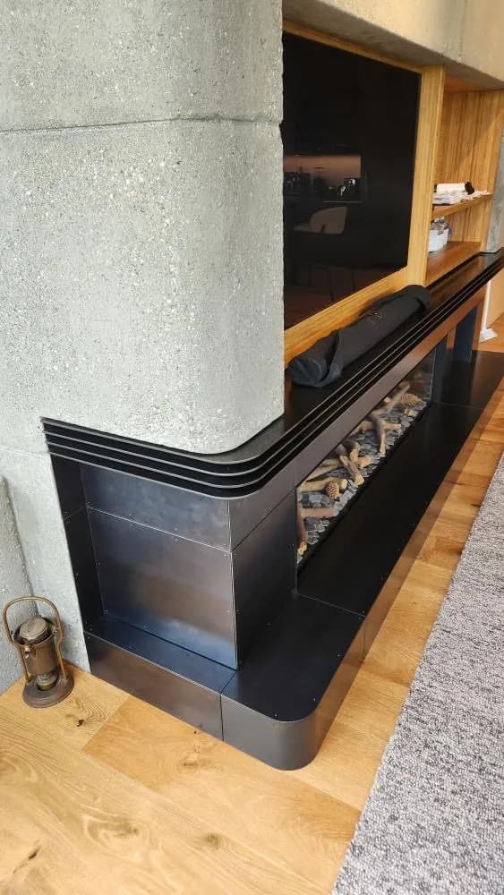 Curved corner of a dark steel fireplace surround, rolled to follow the wall and wrapping around a concrete column