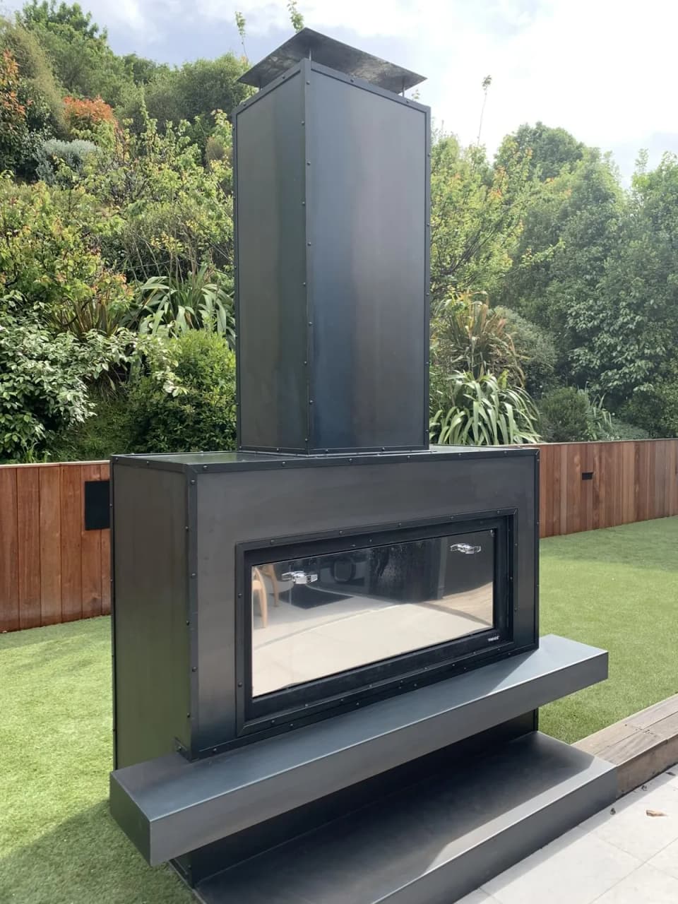 Freestanding black steel outdoor fireplace with a glass front on a lawn