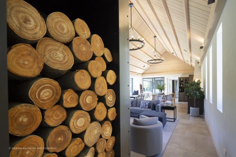 Full-height steel-framed log store wall stacked with firewood beside an open-plan living room