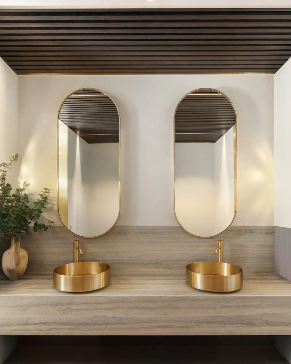 Pair of brass-framed pill-shaped mirrors above twin brass vessel basins on a stone vanity